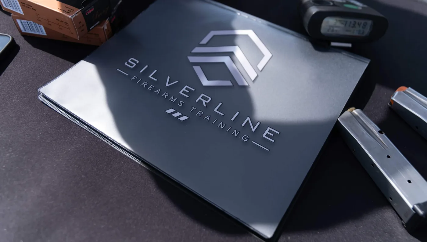 A Silverline Firearms Training binder on the range table, with a shot timer, two pistol magazines and boxes of 9mm ammunition around it.